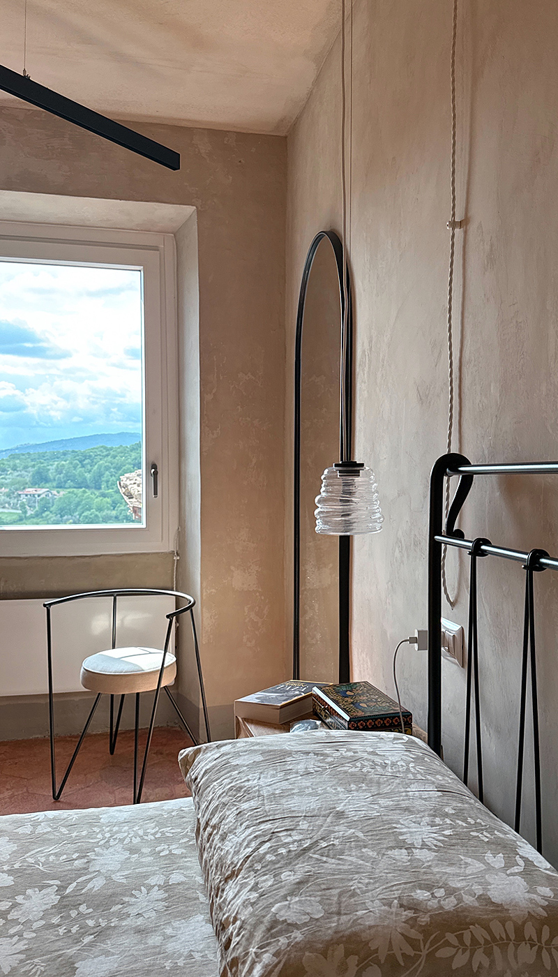 Letto singolo accanto a finestra con panorama collinare, sedia in metallo e lampada ad arco.