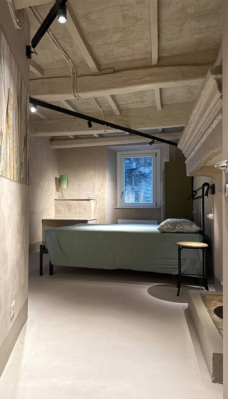 Letto matrimoniale con copriletto verde salvia, accanto a camino e finestra con luce naturale.