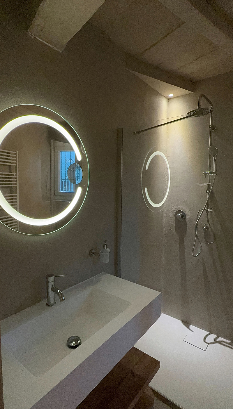 Lavabo rettangolare con specchio rotondo retroilluminato e riflesso sulla parete della doccia.